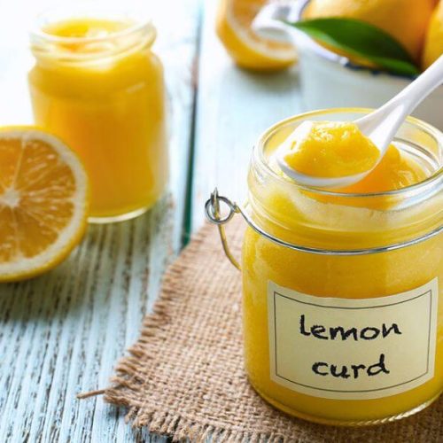 Glass jars of lemon curd with a fresh lemon, lemon zest on a spoon, and rustic wood background—bright and gift-ready.