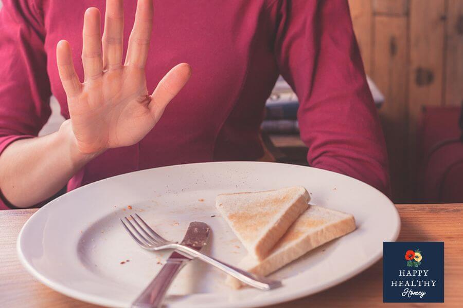 Woman in a red shirt holding her hand up in refusal toward a plate with slices of toast, representing the need to avoid gluten for dietary or medical reasons.