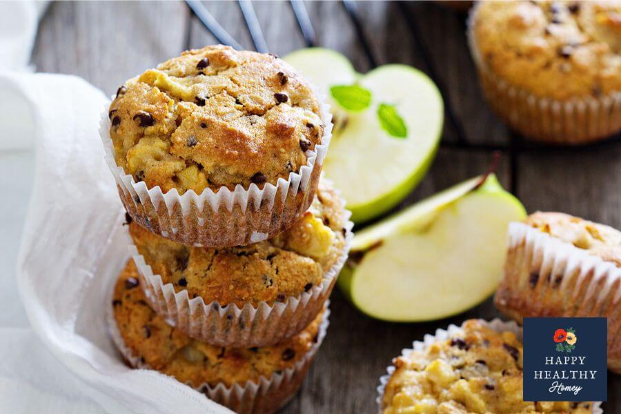 Stack of freshly baked gluten-free apple muffins with mini chocolate chips on a rustic wooden surface, surrounded by sliced green apples and cooling rack.