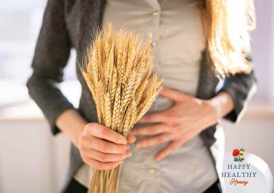 A woman in a gray shirt and black sweater holds a bouquet of wheat with one hand and touches her stomach with the other, symbolizing gluten-related discomfort.