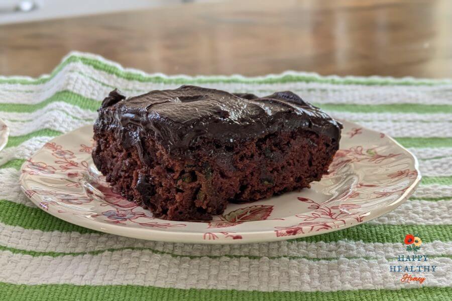 Close-up of a frosted zucchini brownie on a floral-patterned plate, styled on a green and white towel with natural kitchen light.