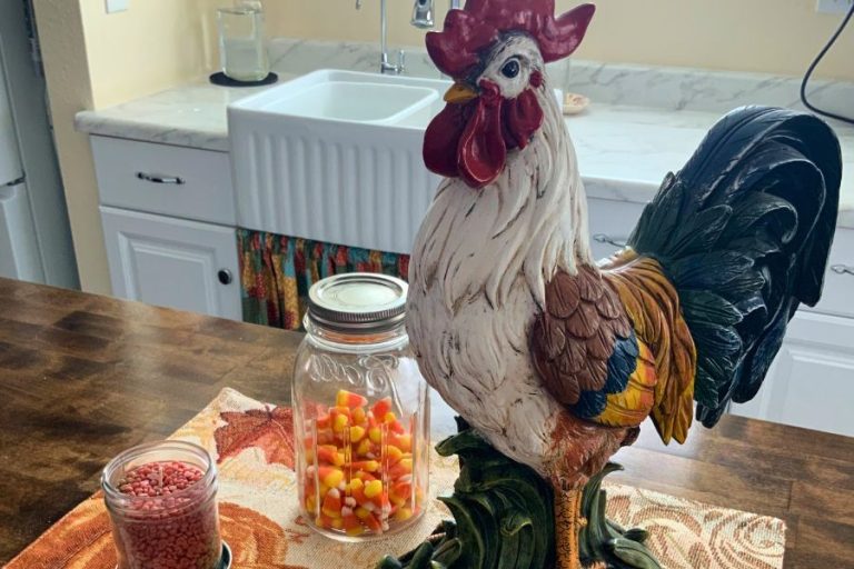 Ceramic rooster fall kitchen centerpiece on a pumpkin-themed placemat with candy corn and autumn decor in a cozy white farmhouse kitchen.