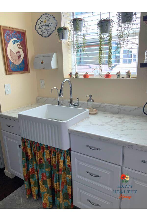 White farmhouse sink with cozy fall sink skirt, pumpkins on the windowsill, and cheerful autumn kitchen decor.