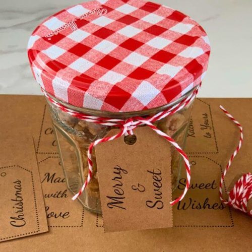 Small jar of homemade candied walnuts with a red gingham lid, twine, and kraft gift tag on top of printable Christmas gift tags.