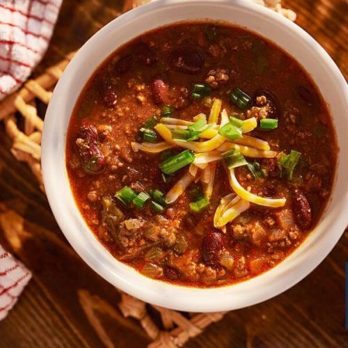 Bowl of chili garnished with green onions and cheese on wooden table with dish towel, and salt and pepper shakers.