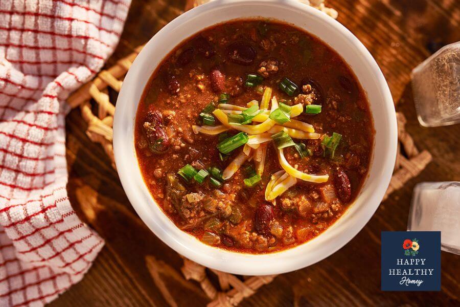 Bowl of chili garnished with green onions and cheese on wooden table with dish towel, and salt and pepper shakers.